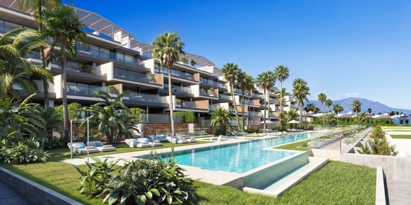 Uitzicht op Nurbay in Manilva, Spanje: moderne witte appartementencomplexen omringd door weelderig groen, met de Middellandse Zee op de achtergrond en een strakke blauwe hemel erboven. Een luxueuze en serene omgeving voor wonen en ontspannen.